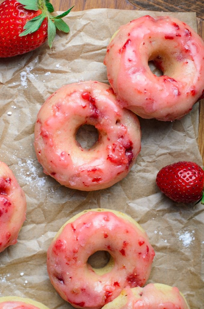 a batch of baked strawberry glazed donuts shown on crinkled brown parchment paper with fresh berries in the background