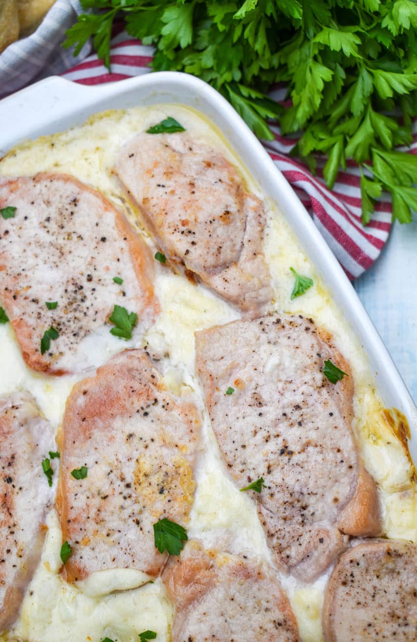 baked pork chop and potatoes in a white casserole dish topped with fresh chopped green herbs