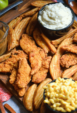 closeup picture of a fried chicken dinner board featuring chicken strips, potato wedges, and bowls of mac and cheese, coleslaw, brown gravy, and creamy mashed potatoes