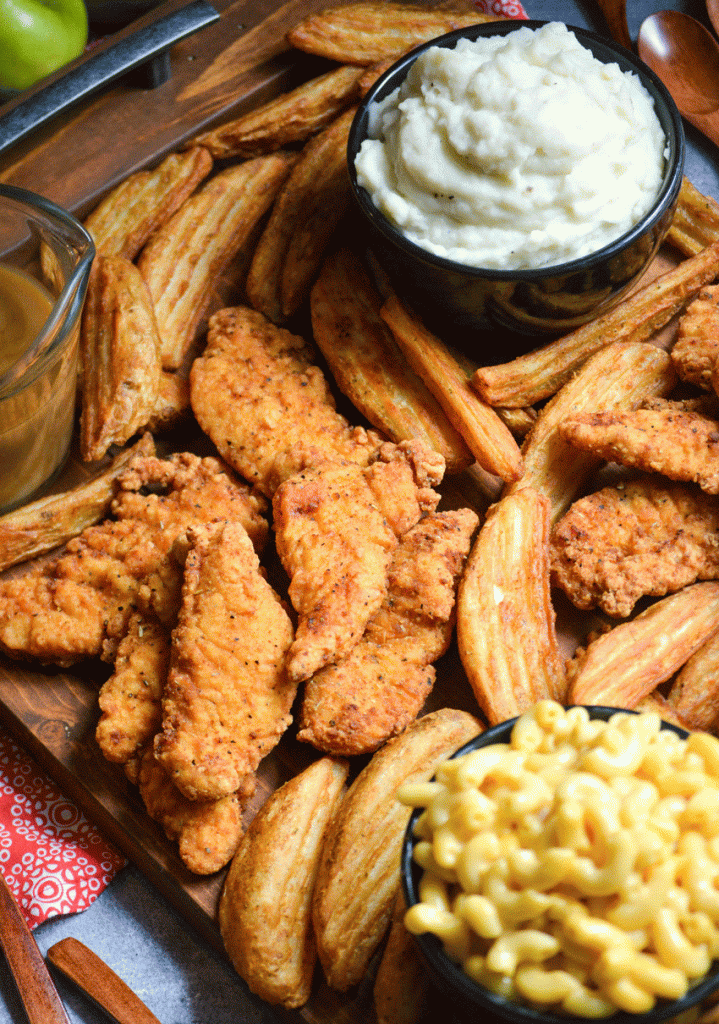 closeup picture of a fried chicken dinner board featuring chicken strips, potato wedges, and bowls of mac and cheese, coleslaw, brown gravy, and creamy mashed potatoes