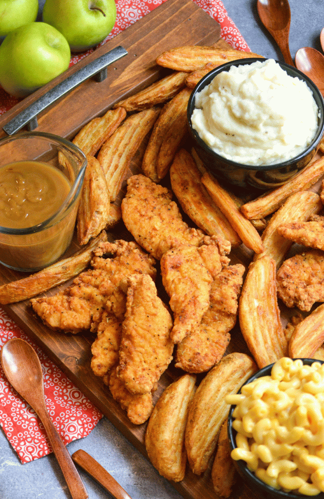 a wooden serving tray covered with fried chicken tenders and crispy potato wedges with bowls of sides in the corners