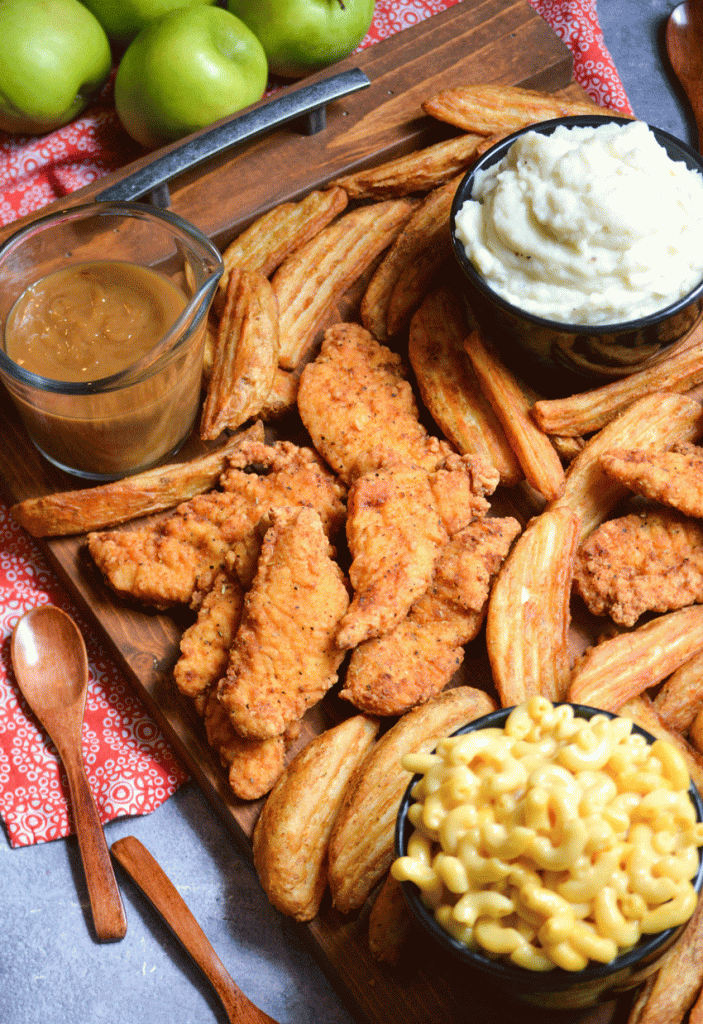 a wooden serving tray covered with fried chicken tenders and crispy potato wedges with bowls of sides in the corners