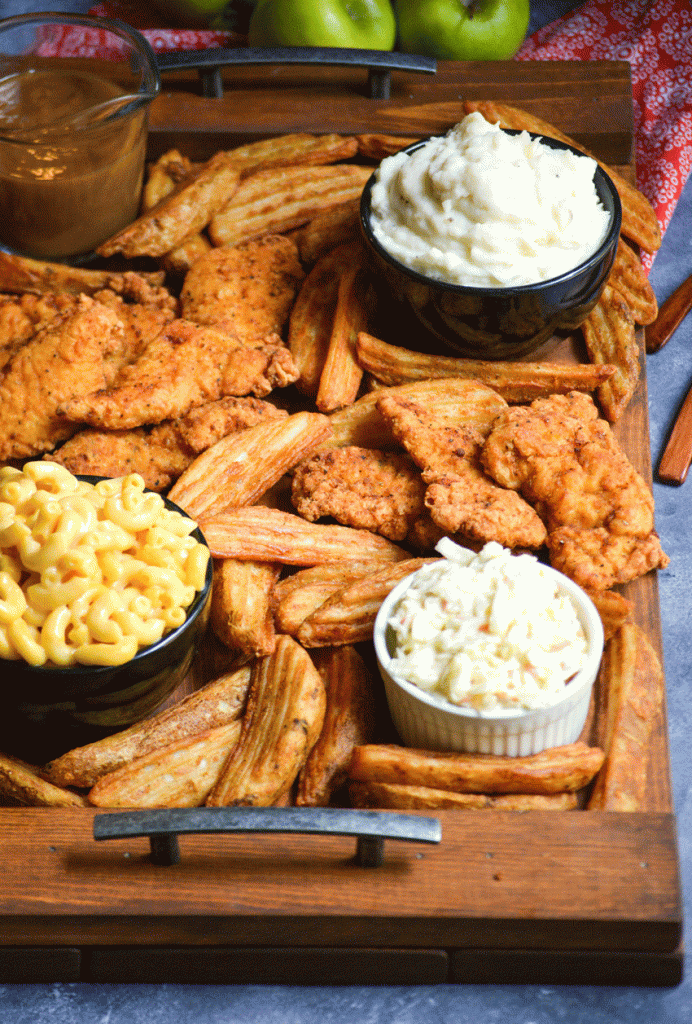 a fried chicken dinner board featuring chicken strips, potato wedges, and bowls of mac and cheese, coleslaw, brown gravy, and creamy mashed potatoes