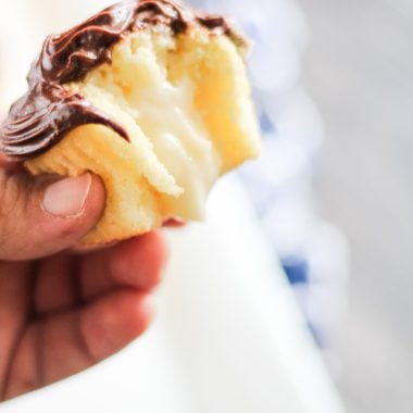 a hand holding up a boston cream pie cupcake with a bite remove to show the cream filling center