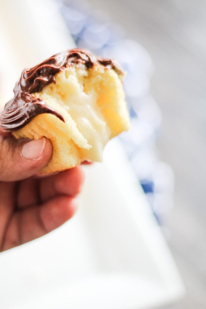 a hand holding up a boston cream pie cupcake with a bite remove to show the cream filling center