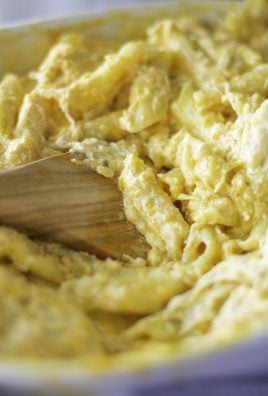 a wooden spatula shown stirring the cheesy baked buffalo chicken mac and cheese together in a white casserole dish