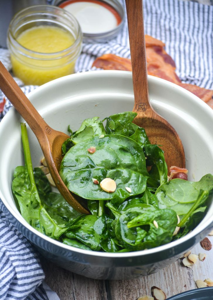 a ceramic mixing bowl filled with lightly dressed spinach salad with wooden utensils sticking out of the salad