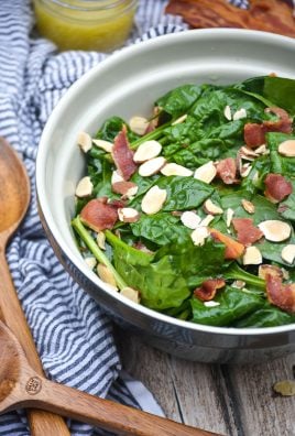 a blue ceramic mixing bowl filled with spinach salad topped with crisp bacon bits and sliced almonds