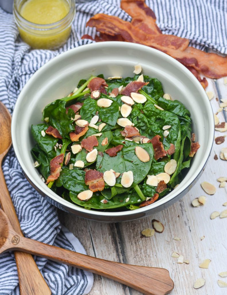 a blue ceramic mixing bowl filled with spinach salad topped with crisp bacon bits and sliced almonds
