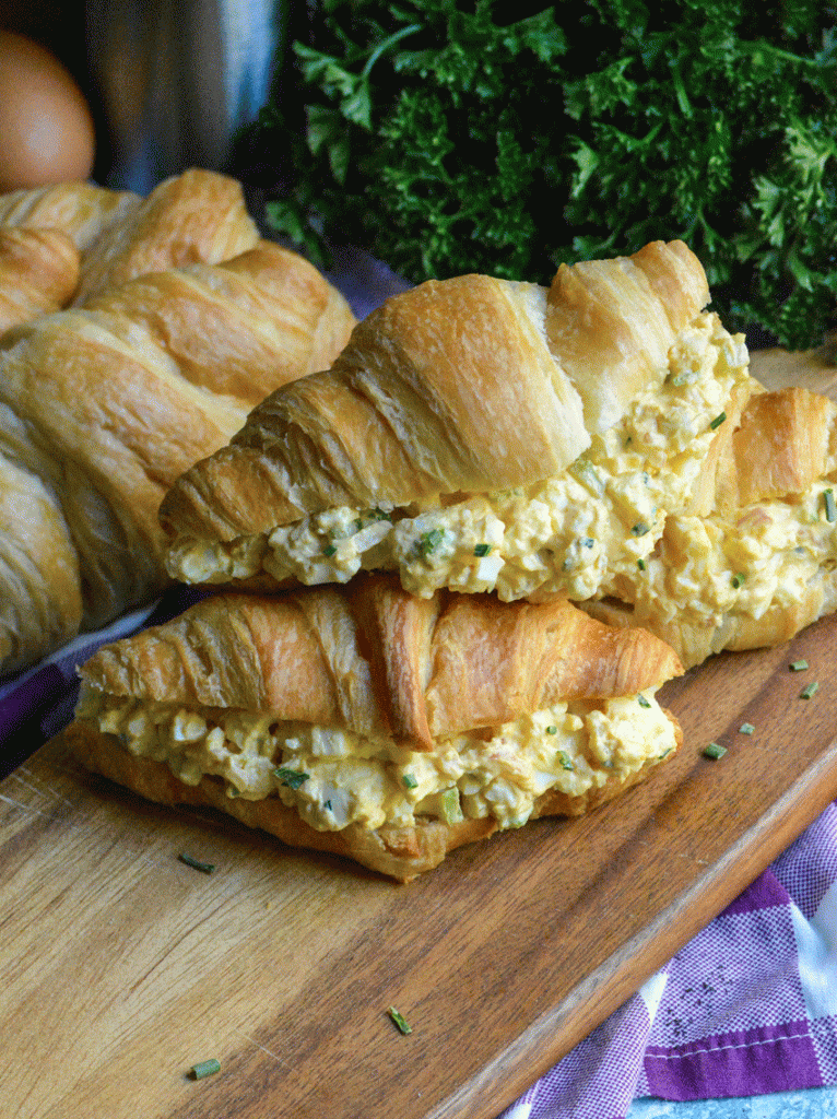 three croissants stuffed with the best egg salad recipe stacked together on a wooden cutting board