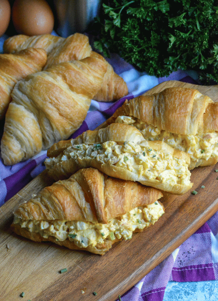 the croissant sandwiches filled with the best egg salad recipe and shown in a single row on a wooden cutting board with the thick filling in full view