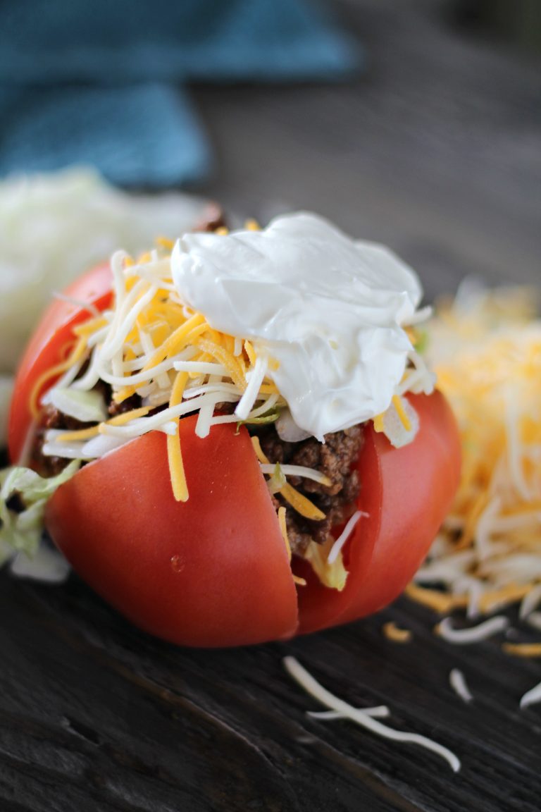 taco stuffed tomato shown on a wooden background and topped with shredded cheeses and a hearty dollop of sour cream