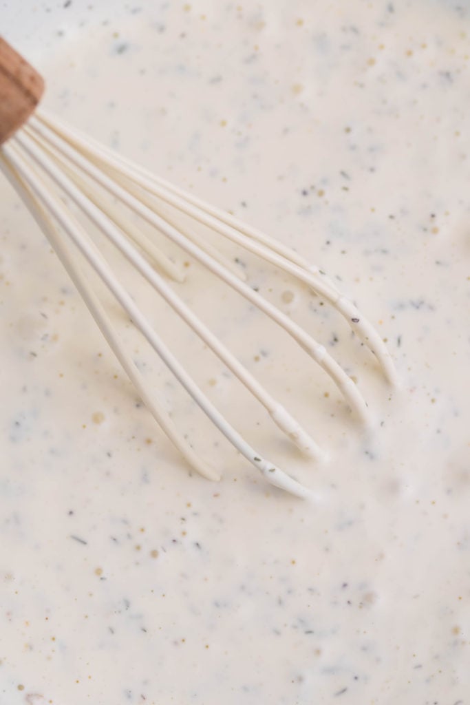 a white whisk shown mixing together homemade ranch dressing in a glass bowl