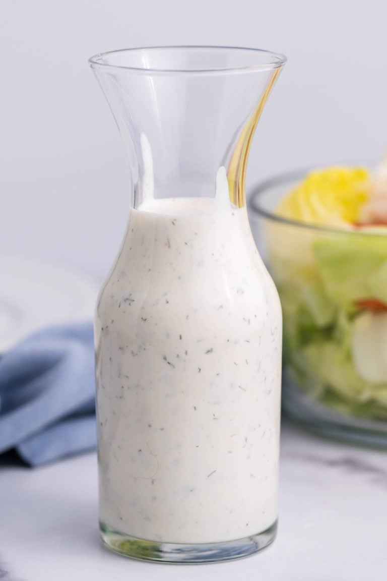 homemade ranch dressing shown in a small glass decanter in front of a glass bowl filled with salad