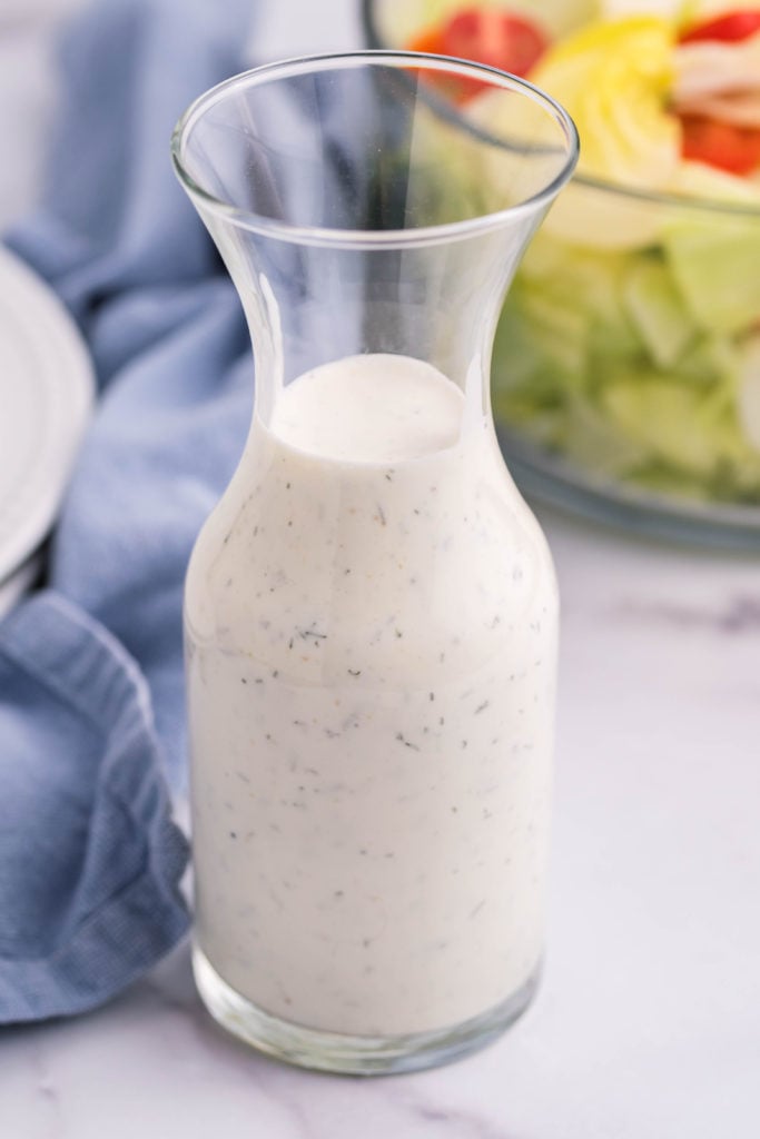 homemade ranch dressing shown in a small glass decanter in front of a glass bowl filled with salad