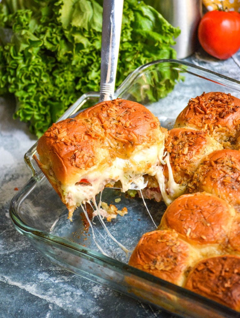 a silver spatula shown lifting two tuna melt sliders out of a glass baking dish with pulled cheese tendrils hanging down