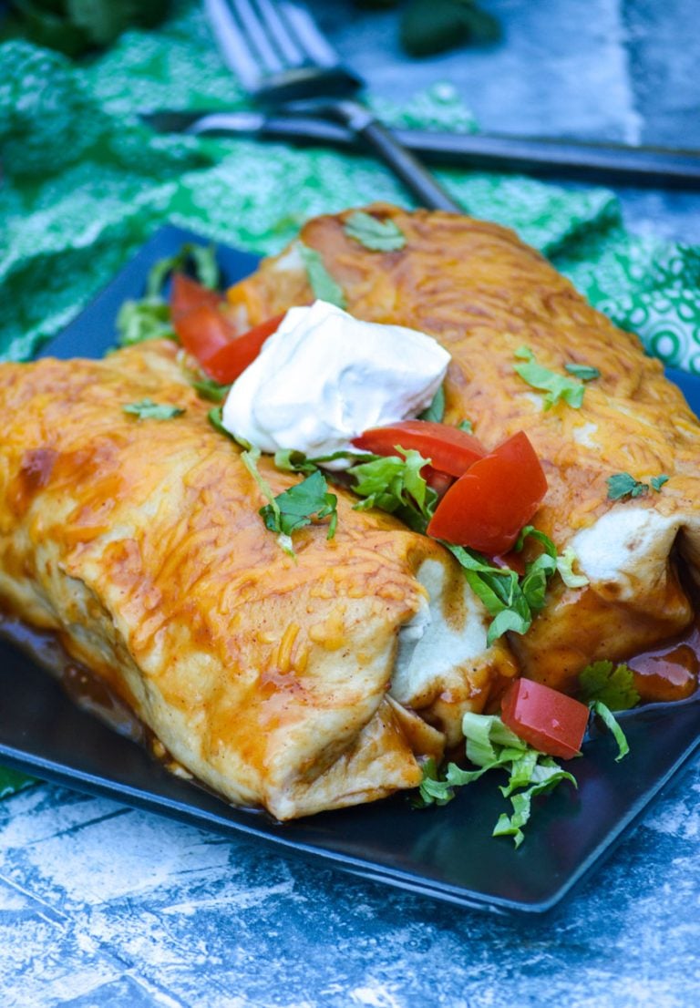 two wet burritos shown on a black square plate topped with cilantro leaves, chopped tomatoes, and sour cream