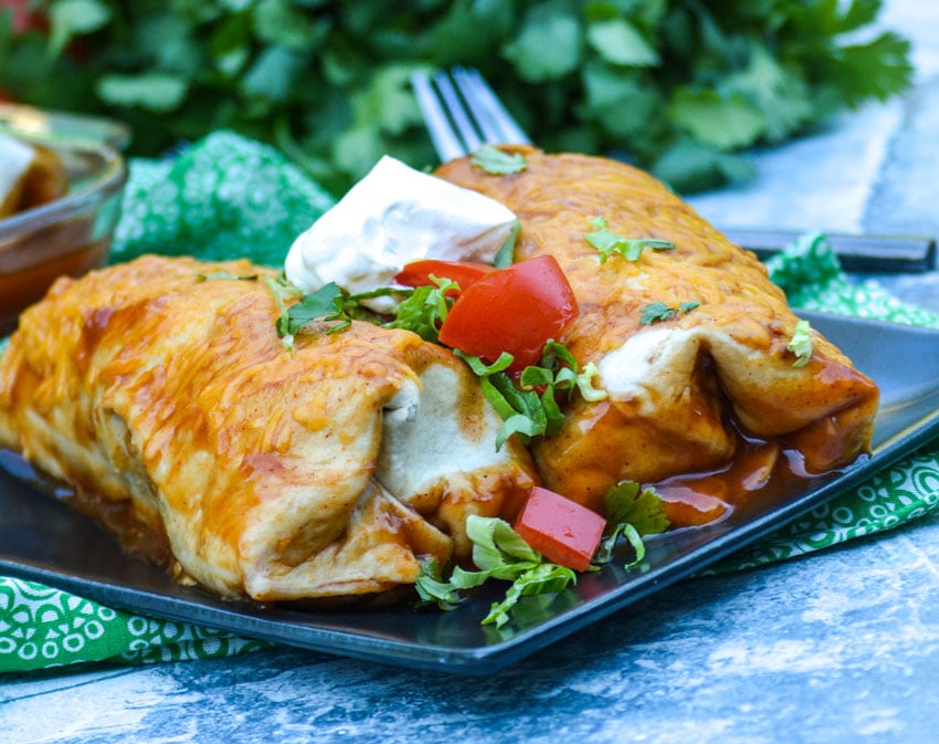 two wet burritos shown on a black square plate topped with cilantro leaves, chopped tomatoes, and sour cream