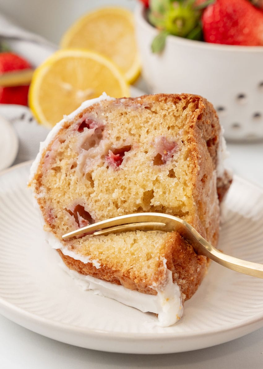 a gold fork slicing a piece of strawberry lemon yogurt cake on a small white plate