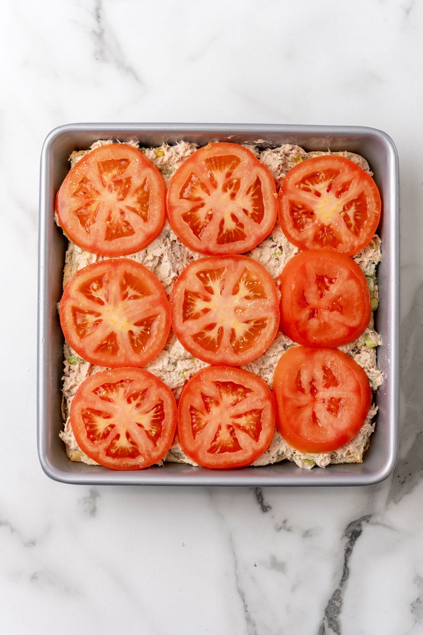 slices of ripe tomatoes overtop tuna salad on slider buns in a square metal baking dish