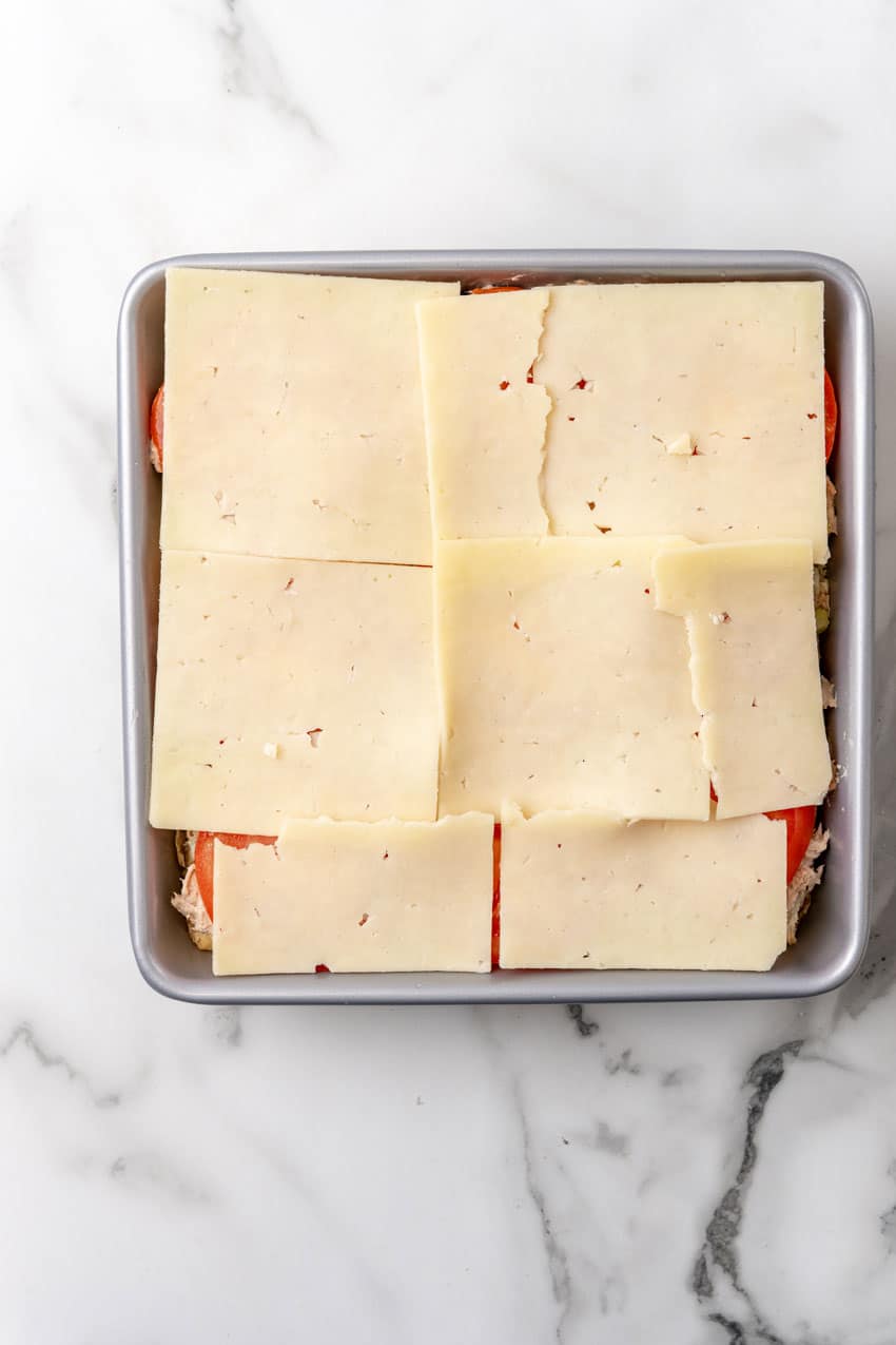 slices of cheese and tomatoes over tuna salad on slider buns in a square metal baking dish