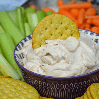 creamy french onion dip shown served in a small purple bowl shown with a cracker stuck in the dip
