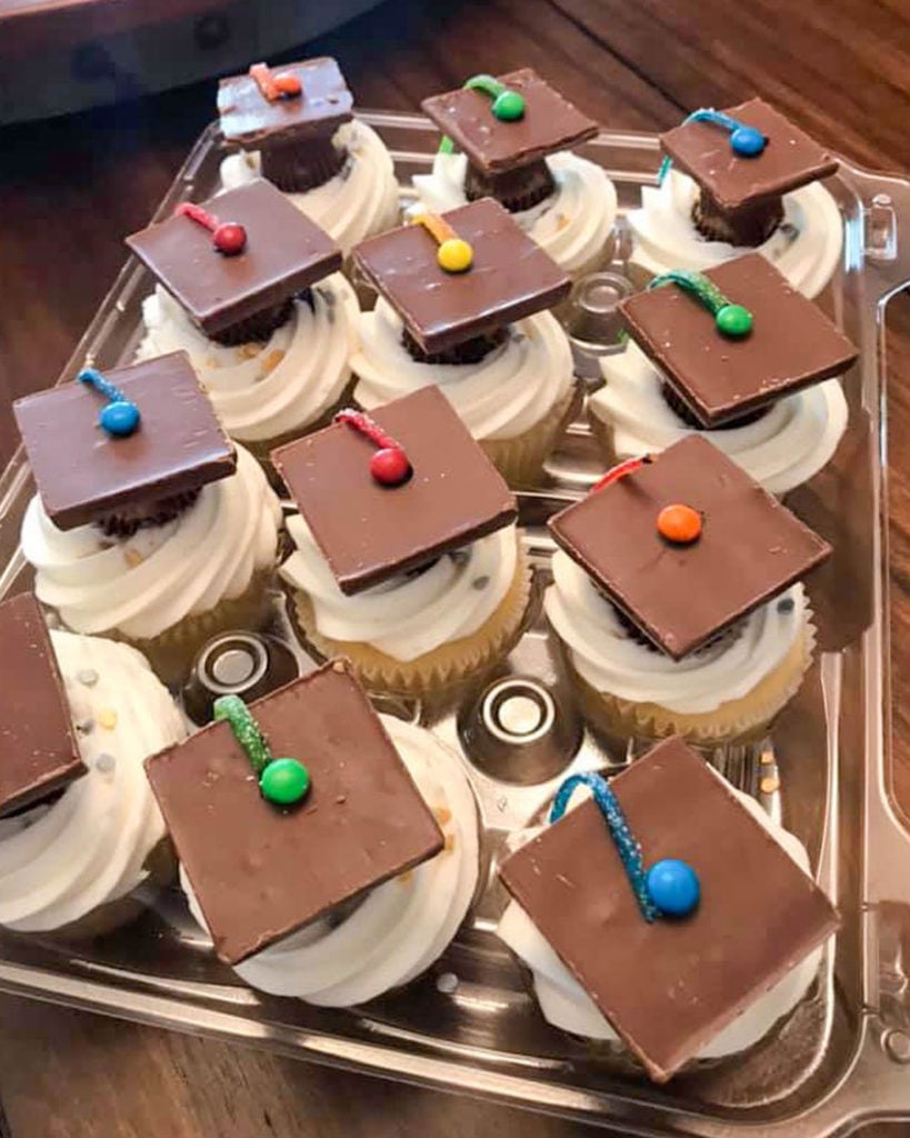 an overhead shot showing graduation cap cupcakes in a clear plastic cupcake holder on a wooden table