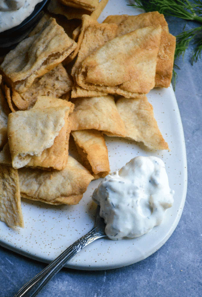 a silver spoon shown resting on a white platter with a scoop of tzatziki sauce and pita chips in the background