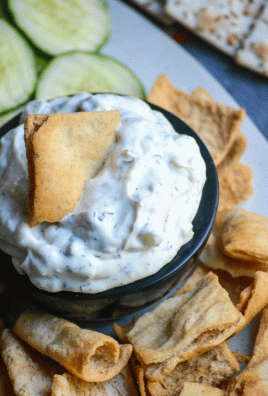a small black bowl filled with homemade tzatziki dip surrounded by cucumber sliced and pita chips