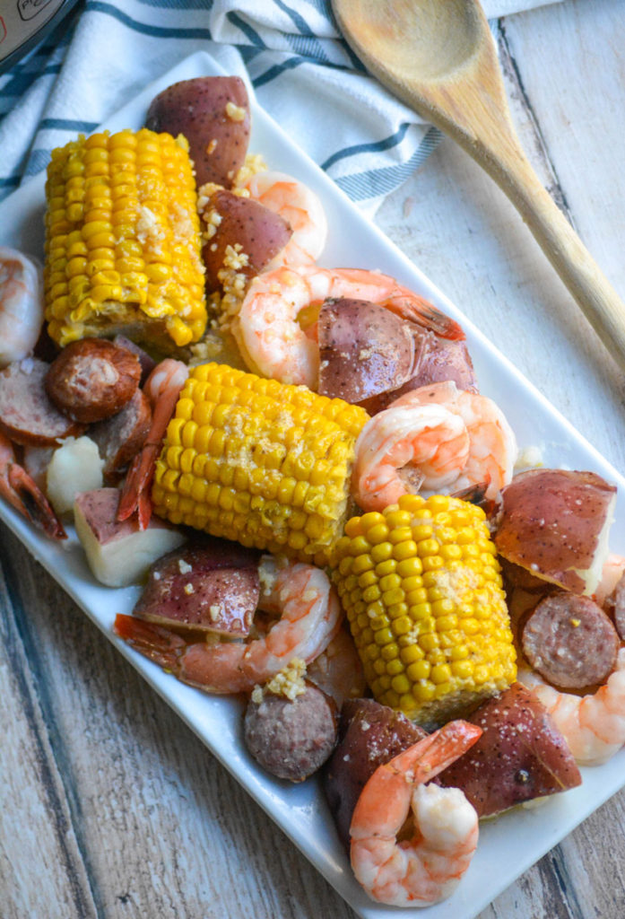 Instant Pot shrimp boil shown on a white platter with a stick of butter, a wooden spoon, and a pressure cooker in the background