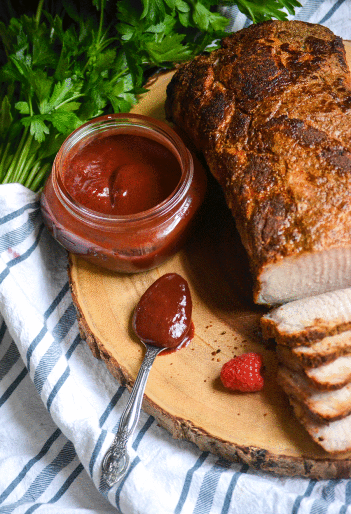 a silver spoon shown full of raspberry dijon barbecue sauce on a wooden cutting board next to a seasoned roasted pork loin with fresh herbs in the background