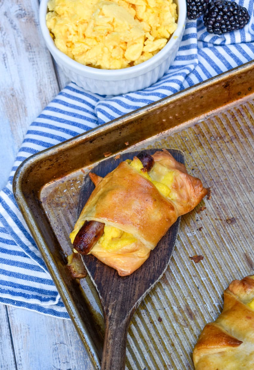 A WOODEN SPATULA LIFTING A BREAKFAST CRESCENT ROLL OFF A METAL SHEET PAN