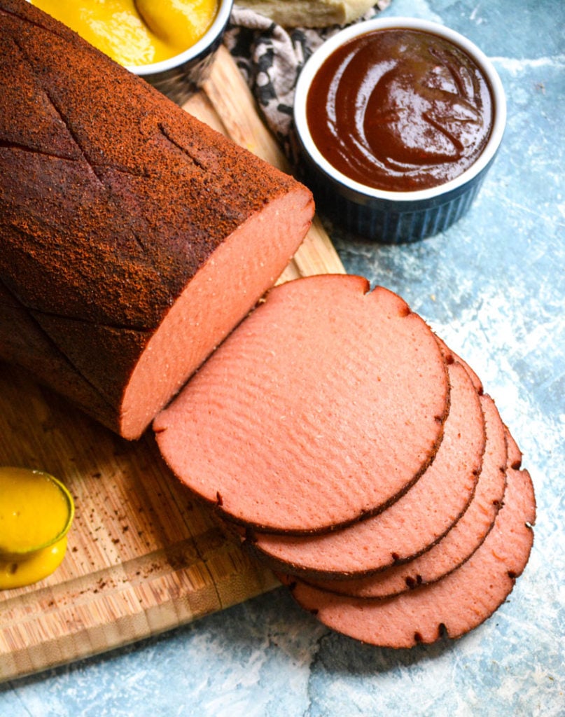 a chub of smoked bologna shown sliced on a wooden cutting board with barbecue sauce & mustard in the background