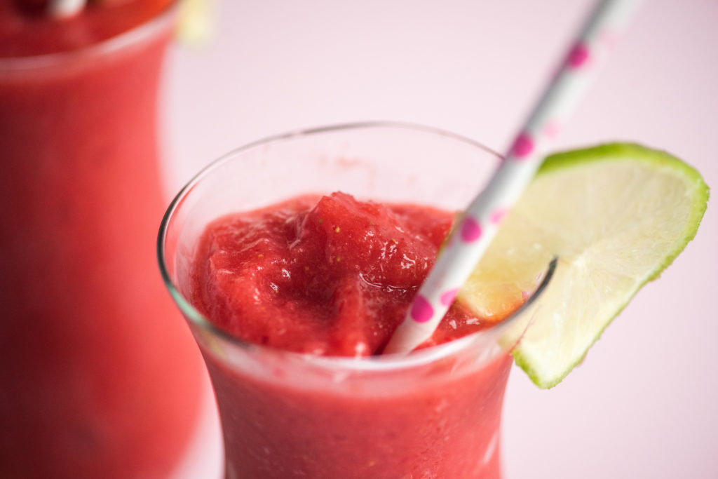 a close up shot of strawberry daiquiris in a glass jar garnished with paper straw and slice of lime