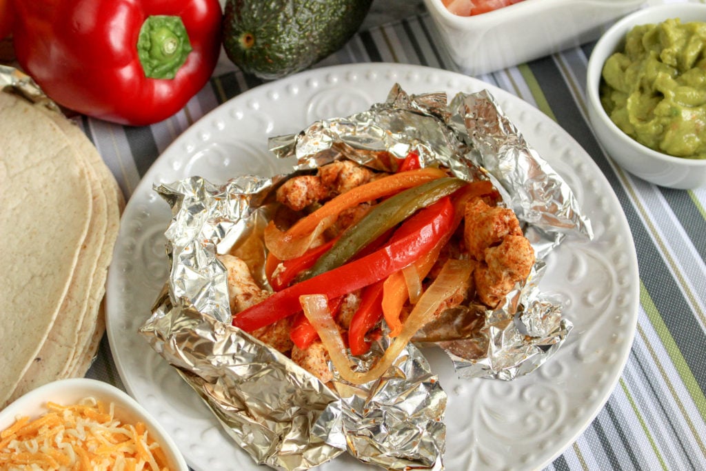 grilled chicken fajita foil packet open on a white plate surrounded by topping options