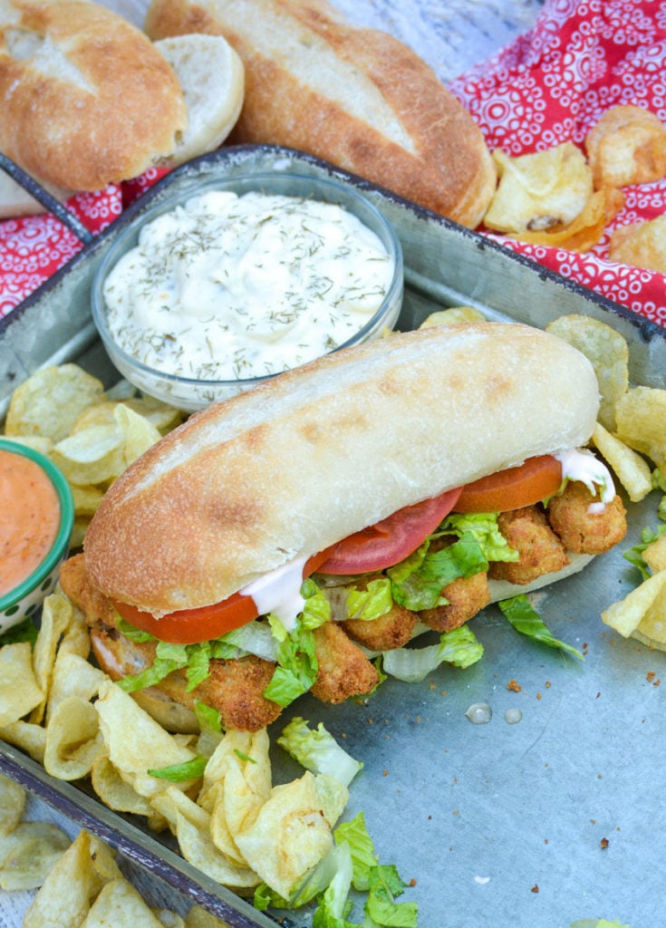 a fish stick po boy sandwich on a metal serving tray with a bowl of tartar sauce for dipping