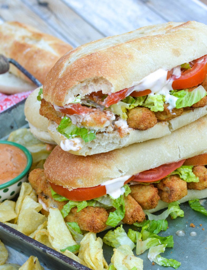 two fish stick po boys stacked together on a metal serving tray surrounded by potato chips