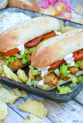 two fish stick po boy sandwiches served on a metal tray with chips and a bowl of tartar sauce