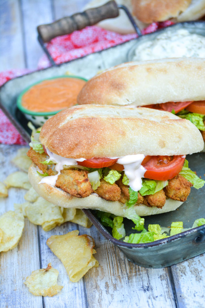 two fish stick po boy sandwiches served on a metal tray with chips and bowls of tartar sauce & remoulade sauce