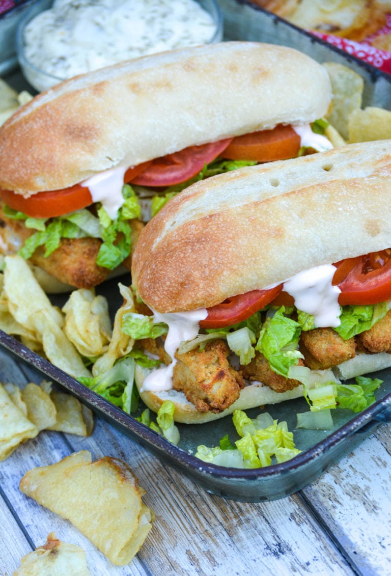 two fish stick po boy sandwiches served on a metal tray with chips and a bowl of tartar sauce