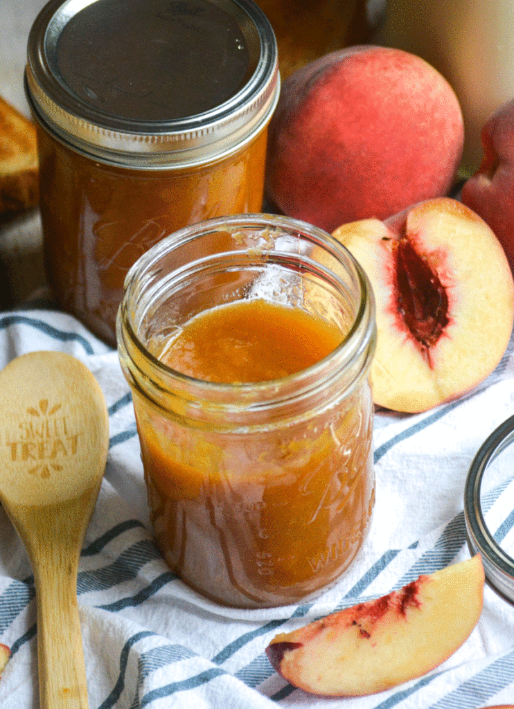 Instant pot peach jam shown in a glass mason jar wit fresh peaches in the background