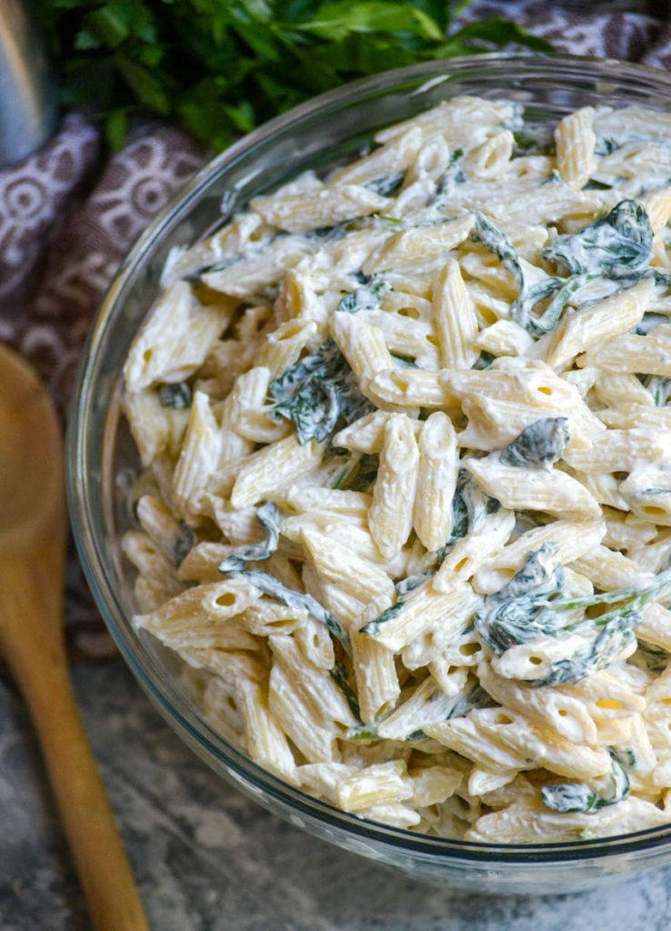 spinach dip pasta salad in a large glass bowl with a wooden spoon on the side