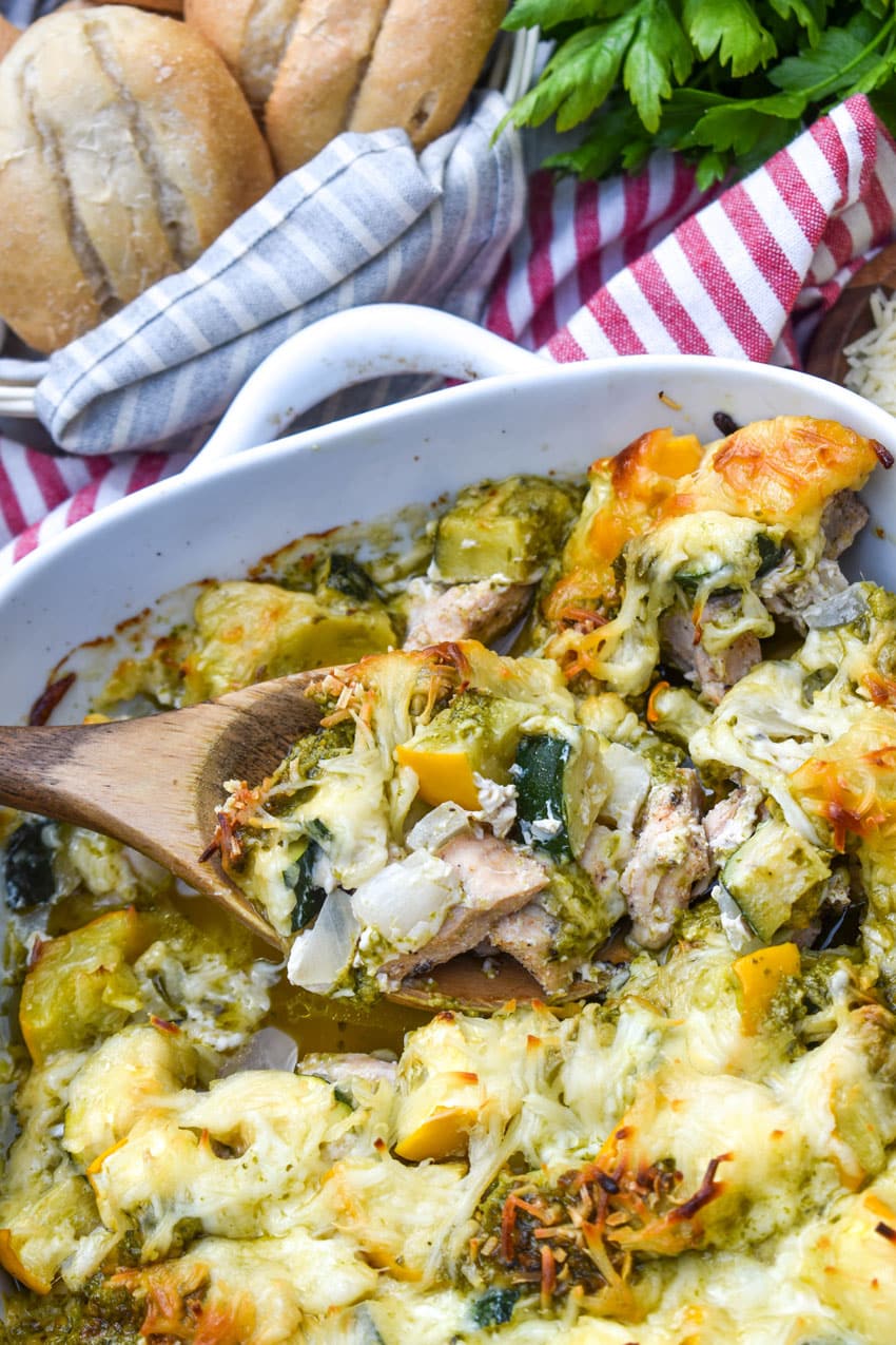 a wooden spoon scooping cheesy pesto chicken and squash out of a white baking dish