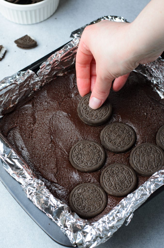 a hand shown laying oreos out evenly over brownie batter in a foil lined baking dish