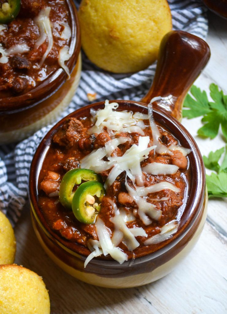 a brown crock style bowl filled with smoked brisket chili garnished with melted cheese and jalapeno slices