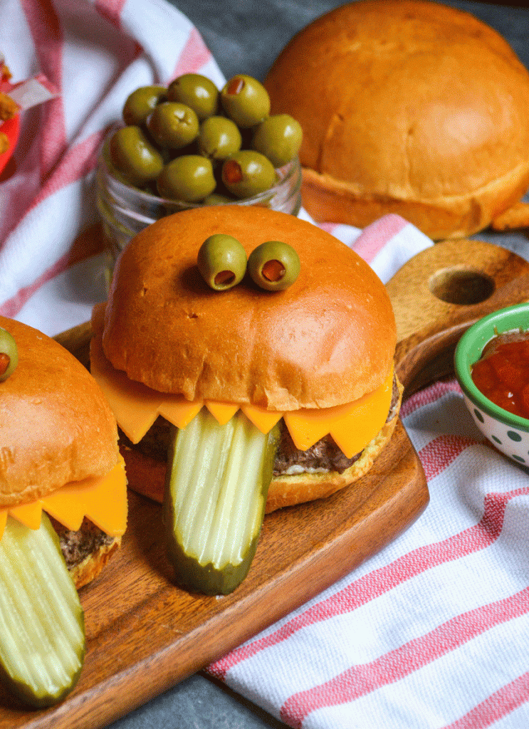 a monster burger with a pickle tongue on a wooden cutting board
