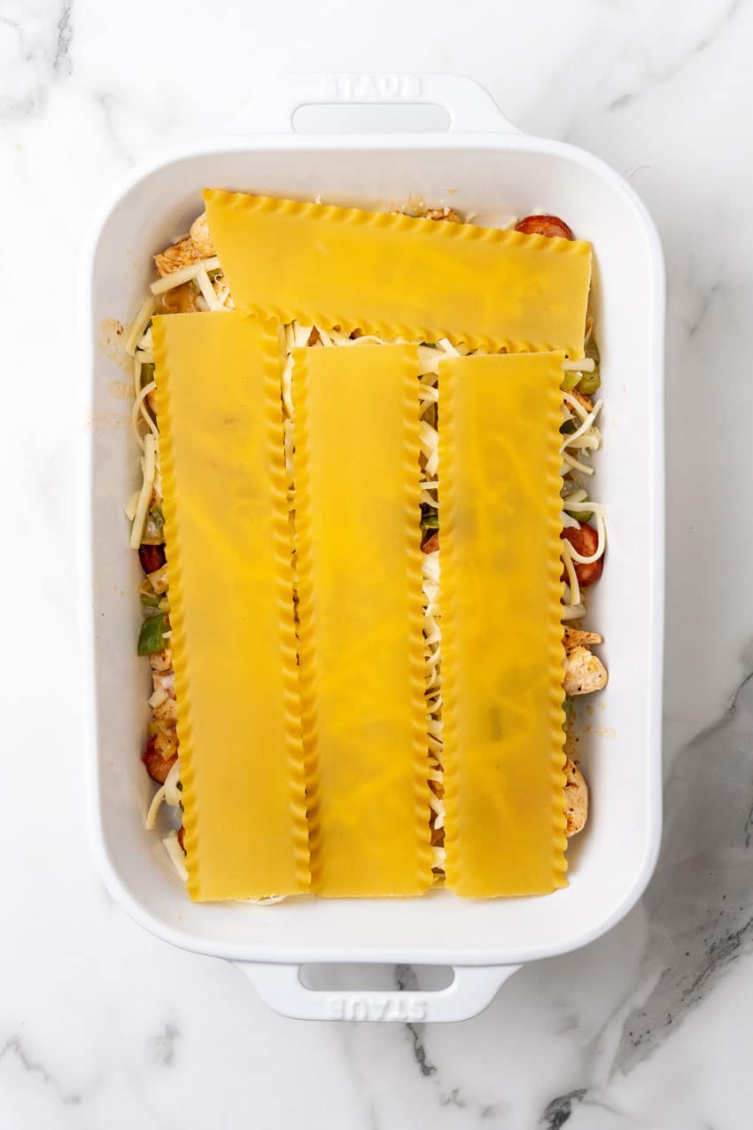 layers of LASAGNA NOODLES TOPPED WITH CAJUN SEASONED MEAT AND VEGGIES and alfredo sauce IN A WHITE BAKING DISH