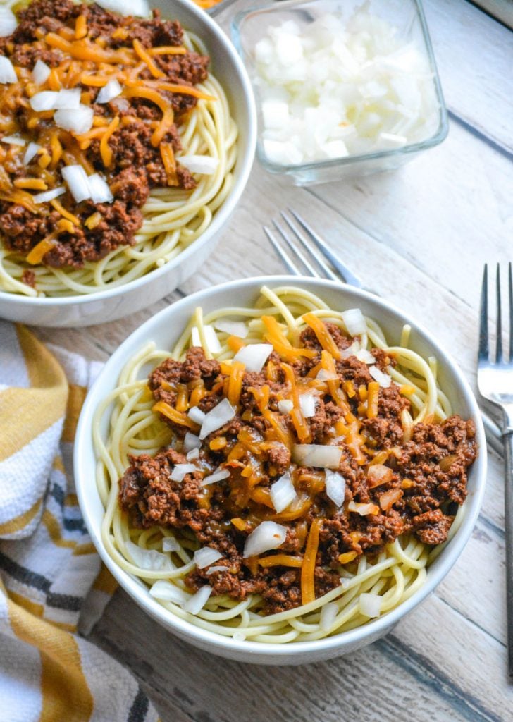 Cincinnati chili served over spaghetti pasta in a white bowl & topped with melted cheddar and diced white onion