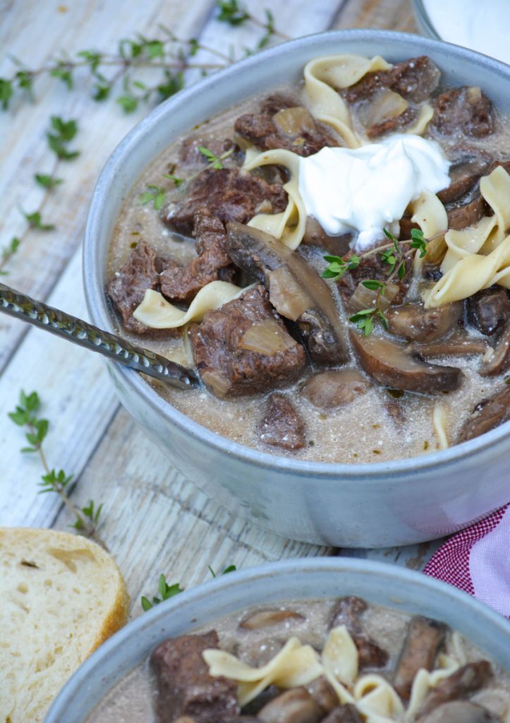 a silver spoon stuck in a bowl of beef stroganoff soup topped with a dollop of sour cream & fresh herbs
