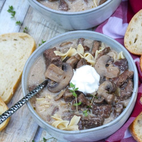 a silver spoon stuck in a bowl of beef stroganoff soup topped with a dollop of sour cream & fresh herbs